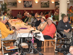Eine Gruppe älterer Menschen sitzt an langen Tischen. Viele tragen auffällige, selbstgestaltete Faschingshüte mit bunten Bändern und Federn. Auf den Tischen stehen Getränke. Einige Personen schauen aufmerksam nach vorne, andere unterhalten sich. Rollatoren stehen neben den Sitzplätzen.