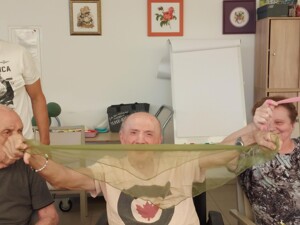 Drei Bewohner:innen sitzen nebeneinander, zwei halten ein langes grünes und ein rosa Tuch gespannt. Ein Schüler steht lächelnd daneben. Die Teilnehmenden wirken aufmerksam und gut in die Aktivität eingebunden.