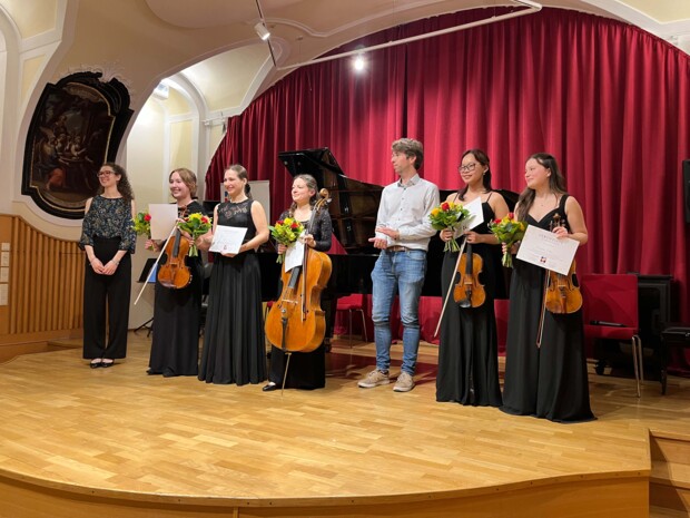 Eine Gruppe junger Musiker:innen steht nebeneinander auf der Bühne, hält Streichinstrumente, Urkunden und Blumensträuße. Im Hintergrund ist ein Flügel vor einem roten Vorhang zu sehen. Eine Person klatscht, während die ausgezeichneten Ensemblemitglieder lächeln.