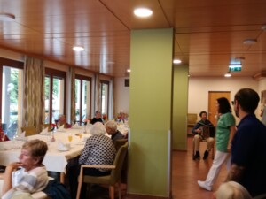 Ein großer Raum mit mehreren gedeckten Tischen, an denen Senior:innen gemeinsam sitzen. Im Hintergrund spielt eine Person Ziehharmonika, während Pflegekräfte sich zwischen den Gästen bewegen. Die Atmosphäre wirkt festlich und freundlich.