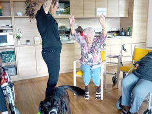 Eine Betreuer:in steht mit erhobenen Armen vor einer sitzenden älteren Person, die dieselbe Bewegung nachahmt. Therapiehund Buddy steht im Vordergrund und blickt direkt in die Kamera.