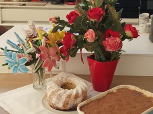 Auf einer Küchenarbeitsplatte stehen ein Gugelhupf mit Staubzucker, eine große Form Tiramisu sowie zwei Blumenarrangements. Im Hintergrund sind eine helle Küchenzeile, Kaffeemaschinen und Geschirr zu sehen.