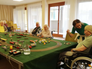 Mehrere Senior:innen sitzen um einen großen Tisch mit einer Modelleisenbahnanlage. Eine Betreuerin spricht freundlich mit einem Mann im Rollstuhl, während andere Teilnehmende sich auf die Züge und Miniaturen konzentrieren. Die Atmosphäre wirkt lebendig und einladend.