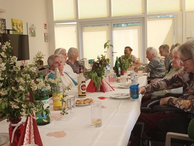 Bewohner:innen sitzen an einem langen, weiß gedeckten Tisch im hellen Gemeinschaftsraum. Der Tisch ist mit Blumen, roten Herzen, Getränken, Gläsern und Mehlspeisen dekoriert. Die Gruppe verbringt einen geselligen Nachmittag in vertrauter Atmosphäre.
