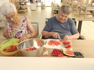 Zwei Bewohner:innen schneiden an einem Tisch Erdbeeren und Wassermelone für eine Sommerbowle. Auf dem Tisch stehen Schalen mit Beeren, Melonenstücken und eine große Metallschüssel.