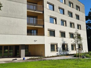 Frontansicht des neuen Pflegewohnhauses mit klarer, moderner Architektur. Vor dem Gebäude liegt eine große grüne Rasenfläche mit neu gepflanzten Bäumen.
