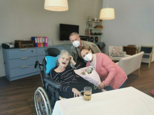 Bewohnerin an Tisch sitzend im neuen ÖJAB-Pflegewohnhaus, daneben Angehörige stehend mit Blumenstrauß in der Hand.