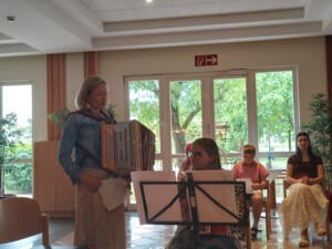 Elisabeth Unger steht mit einer Ziehharmonika neben einer Schülerin, die hinter einem Notenständer spielt. Weitere Schüler:innen sitzen im Hintergrund, der Raum ist hell und freundlich.