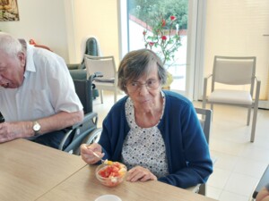 Eine ältere Bewohnerin sitzt mit einer Schüssel Obstsalat am Tisch und isst. Neben ihr probiert ein älterer Bewohner ebenfalls Obstsalat; im hellen Aufenthaltsraum stehen Rollstühle, Stühle und Pflanzen.
