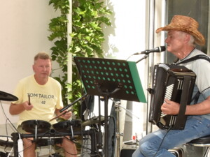 Ein Mann spielt Schlagzeug, daneben sitzt ein Musiker mit Akkordeon und singt ins Mikrofon. Beide musizieren vor einem Notenständer, die Sonne scheint in den Garten.