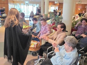 Mehrere Kinder und ältere Personen in Rollstühlen lachen und unterhalten sich während der Übergabe von Geschenkkörben. Die Atmosphäre ist lebendig und herzlich.