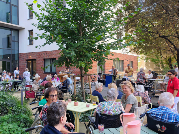 Mehrere Senior:innen sitzen in geselliger Runde an Gartentischen im begrünten Innenhof des ÖJAB-Pflegewohnhauses Neumargareten. Pfleger:innen und Besucher:innen unterhalten sich mit den Bewohner:innen, während im Hintergrund Musik gespielt wird. Auf den Tischen stehen Gläser mit Bowle, Teelichter und Krüge.
