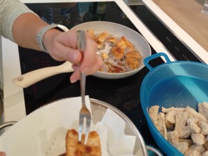 Eine Person hebt mit zwei Gabeln frisch gebackene panierte Steinpilze aus einer Pfanne in eine Schüssel mit Küchenpapier. Im Hintergrund stehen Ölflasche und Wasserflaschen.