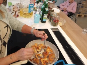 Eine Person brät panierte Steinpilze in einer Pfanne. Auf dem Tisch daneben stehen Öl, Wasserflaschen und Dekogläser. Im Hintergrund beobachten zwei ältere Bewohner:innen das Geschehen.