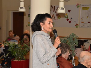 Eine Frau mit dunklem, zusammengebundenem Haar spricht in ein Mikrofon. Sie trägt einen grauen Pullover und steht zwischen den Gästen der Feier.