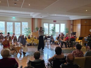 Ein Raum voller Bewohner:innen, viele davon in Rollstühlen, schaut aufmerksam auf eine Bühne. Dort spielen Kinder ein Krippenspiel, begleitet von musikalischen Beiträgen. Eine erwachsene Person spricht ins Mikrofon.