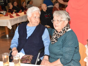 Zwei Frauen - eine Seniorin und ihre Angehörige - sitzen nebeneinander an einem Tisch. Im Hintergrund sind weitere Gäste in einem Gemeinschaftsraum zu sehen.