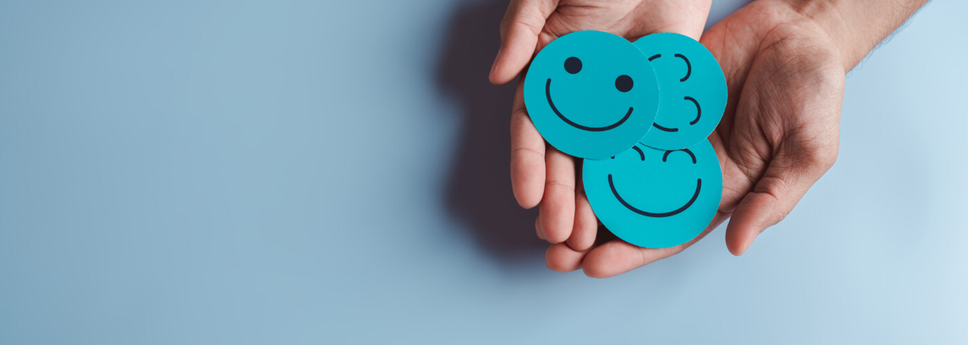Symbolbild. Zwei geöffnete Hände halten drei türkisfarbene runde Karten mit verschiedenen Smileys, die Freude und positive Emotionen ausdrücken. Der Hintergrund ist einfarbig hellblau.
