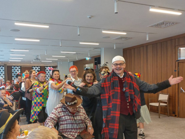 Bewohner:innen und Mitarbeiter:innen, zu Fasching fröhlich verkleidet, bei einer Polonaise im Festsaal des Hauses.