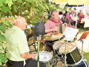 Die Musiker Rudi und Ernstl sitzend beim Musizieren am Frühlingsfest für BewohnerInnen. Im Hintergrund BewohnerInnen sitzend.