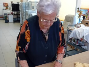Eine Seniorin formt einen langen Teigstrang auf einem mit Mehl bestäubten Holztisch. Neben ihr liegen Teigstücke bereit.