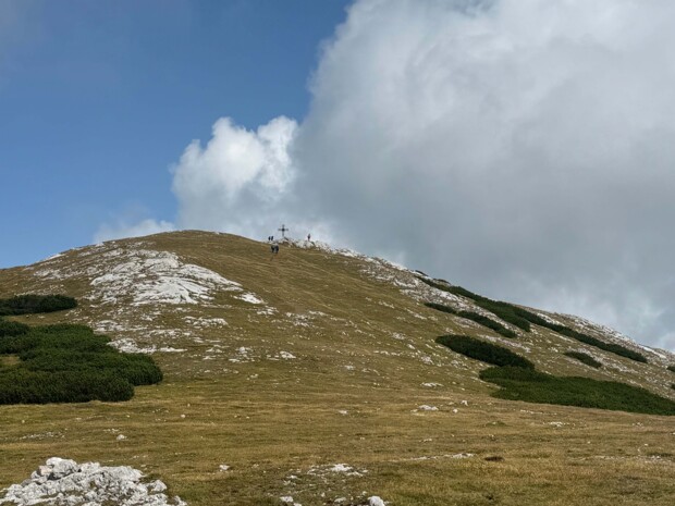 Ein leicht ansteigender, kahler Berghang mit einzelnen Sträuchern und Felsflächen. In der Ferne ist das Gipfelkreuz zu erkennen, mehrere Personen sind auf dem Weg dorthin. Der Himmel ist teils wolkig.