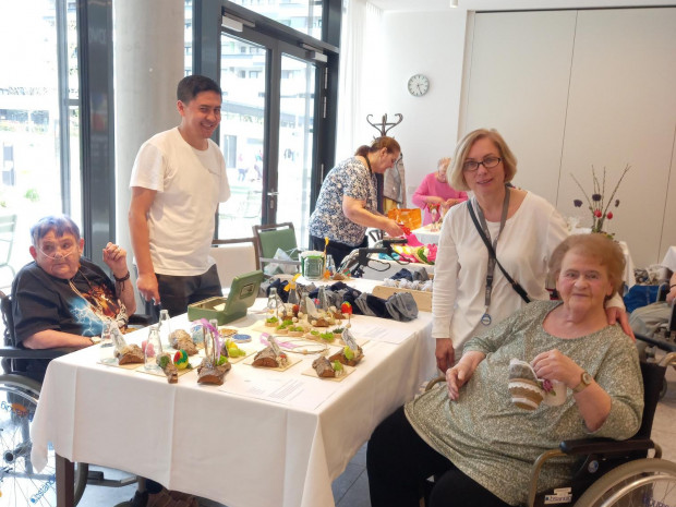 Eine Gruppe von Personen, darunter mehrere Rollstuhlnutzer:innen, steht und sitzt um einen mit handgefertigter Osterdekoration gedeckten Tisch. Alle wirken fröhlich und im Gespräch vertieft.