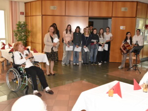 Eine ältere Frau im Rollstuhl spricht in ein Mikrofon, neben ihr steht eine Lehrkraft. Im Hintergrund: eine Gruppe Jugendlicher sowie Musiker:innen.