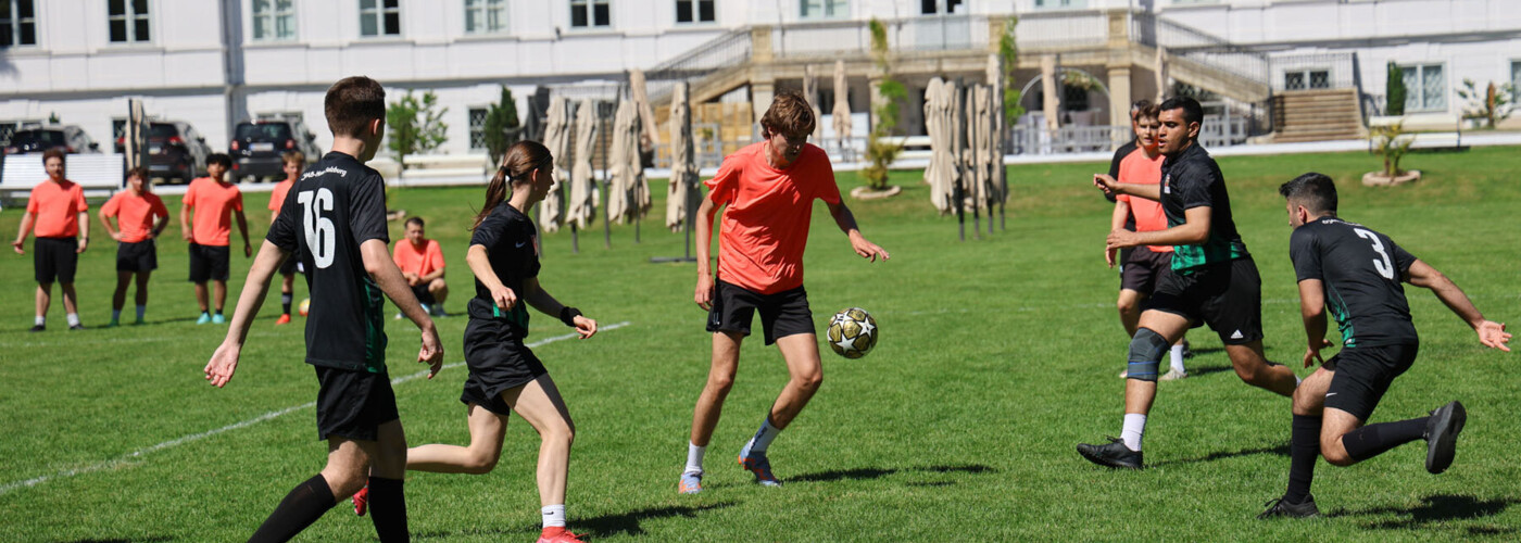 Mehrere Fußballspieler:innen in schwarzen und orangen Trikots laufen auf einer grünen Wiese dem Ball nach. Im Hintergrund steht ein großes weißes Gebäude mit rotem Dach; daneben sind Sonnenschirme, Bäume und Zuseher:innen zu sehen.