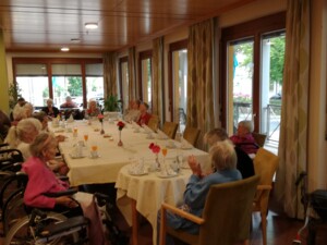 Senior:innen sitzen rund um einen langen, festlich gedeckten Tisch mit Getränken, Blumen und Servietten. Einige Personen applaudieren, andere unterhalten sich. Durch die Fenster fällt Tageslicht, draußen blühen Blumen. Die Stimmung wirkt gelassen und feierlich.
