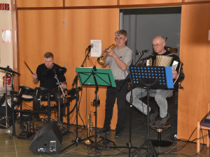 Drei Musiker spielen Trompete, Akkordeon und Schlagzeug auf einer kleinen Bühne im Veranstaltungsraum des Heims.