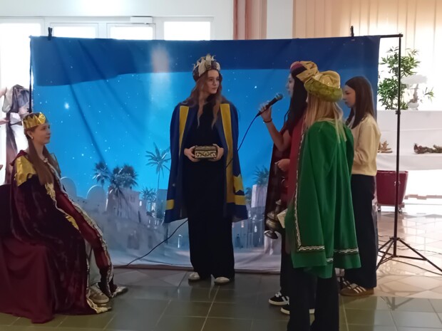 Vier Jugendliche in farbenfrohen Gewändern stellen die Heiligen Drei Könige dar und überreichen Geschenke. Eine Person sitzt mit Krone auf einem Stuhl, drei weitere stehen davor, eine davon spricht ins Mikrofon. Im Hintergrund ein Bühnenbild mit Nachthimmel und Palmen.