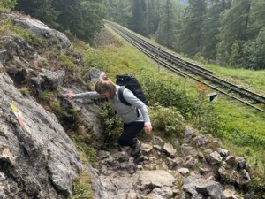 Eine Person mit Rucksack erklimmt vorsichtig einen felsigen Abschnitt eines Wanderwegs. Rechts daneben verlaufen die Gleise der Schneebergbahn. Der Weg ist mit Markierungen versehen, der Bereich ist bewachsen und waldnah.