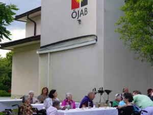 Vor der Fassade des ÖJAB-Hauses St. Franziskus sitzen Bewohner:innen, Angehörige und Mitarbeiter:innen an langen, weiß gedeckten Tischen im Freien. Mehrere Rollstühle und Rollatoren stehen neben den Tischen, auf denen Getränke und kleine Dekorationen stehen.