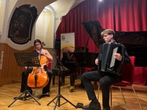Zwei Musiker:innen sitzen auf der Bühne und musizieren gemeinsam. Eine Person spielt Violoncello, die andere Akkordeon. Notenständer stehen davor, im Hintergrund ist ein Flügel und ein roter Vorhang sichtbar.