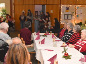 Eine Gruppe Jugendlicher steht nebeneinander und singt Weihnachtslieder. Zwei von ihnen begleiten die Lieder auf Harmonika und Gitarre. Ältere Zuhörer:innen sitzen an einem langen Tisch mit weihnachtlicher Dekoration.