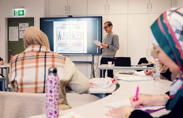 Ein Kursleiter steht mit einem Blatt Papier neben einem großen Bildschirm, auf dem eine Präsentationsfolie zum Thema Aktiv und Passiv zu sehen ist. Kursteilnehmende sitzen an Tischen, schreiben mit und verfolgen den Unterricht. Foto: www.inframe.at