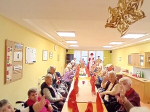 Ein großer Raum voller Bewohner:innen, die an einer langen festlich gedeckten Tafel sitzen. In der Mitte der Tafel stehen rote Geschenksäckchen. Der Nikolaus steht am Ende des Tisches, umringt von fröhlichen Menschen.