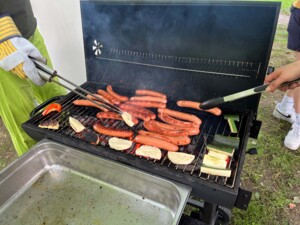 Nahaufnahme eines Grills mit einer Vielzahl an Würstchen und Gemüsestücken. Zwei Personen wenden das Grillgut mit Zangen. Rauch steigt auf, im Hintergrund sind Beine weiterer Personen zu sehen.