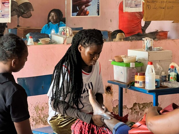 In einem kleinen Salon pflegt eine junge Frau konzentriert den Fuß einer sitzenden Kundin. Zwei weitere Personen stehen daneben und beobachten den Vorgang. Auf einem Tisch liegen Pflegeprodukte und Arbeitsmaterialien, an der Wand hängen Spiegel und Beispielbilder für Kosmetik- und Nageldesign. Die Szene zeigt handwerkliche Dienstleistung und Lernen im Rahmen des beruflichen Alltags in einem einfachen Innenraum.