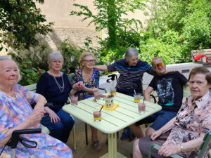 Sechs Senior:innen und ein jüngerer Besucher sitzen entspannt um einen Gartentisch, auf dem Gläser mit Bowle und ein dekorierter Becher stehen. Alle lachen oder lächeln. Im Hintergrund sitzt eine weitere Seniorin auf einer Bank und schreibt an einem Tisch. Der Garten ist grün und blühend.