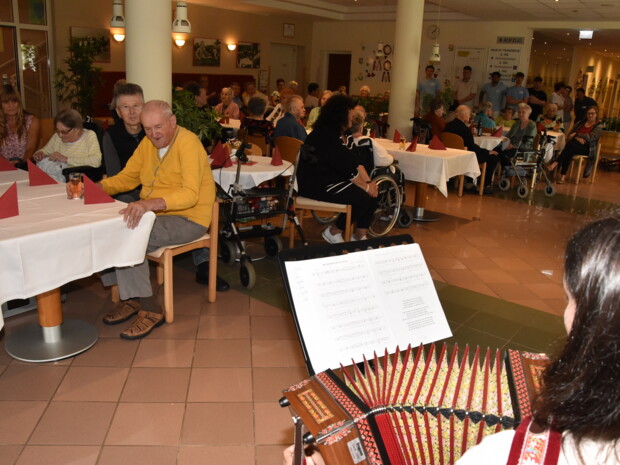 Zahlreiche Bewohner:innen und Gäste sitzen an festlich gedeckten Tischen und lauschen der Musik. Im Vordergrund ist ein Notenblatt und eine Steirische Harmonika zu sehen.