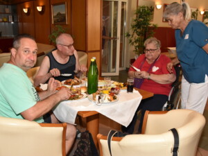 Drei Personen sitzen in der Eingangshalle an einem Tisch und essen gegrillte Speisen. Eine Mitarbeiterin steht daneben und unterhält sich mit den Gästen. Auf dem Tisch stehen gefüllte Teller, Gläser und eine große Flasche Wasser.