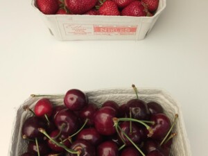 Zwei Pappschalen mit frischem Obst stehen auf einem hellen Tisch. Oben liegen rote Erdbeeren, darunter dunkle Kirschen mit Stielen.