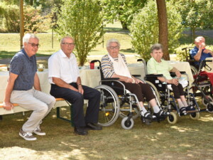 Mehrere Personen, darunter viele im Rollstuhl, sitzen an langen Tischen im Freien. Blaue Sonnenschirme spenden Schatten, einige unterhalten sich lebhaft.