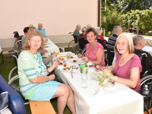 Eine Seniorin im Rollstuhl sitzt mit zwei Angehörigen an einem Gartentisch. Auf den Tellern liegen verschiedene Grillgerichte und Salate, im Hintergrund sind weitere Gäste im Garten zu sehen.