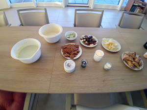 Ein Tisch mit zwei Schüsseln, gefüllt mit Gurke-Topfen-Mischung und anderen Pflegezutaten, sowie Tellern mit Chips, Pralinen und Gebäck. Mehrere Duftkerzen sorgen für eine angenehme Atmosphäre.