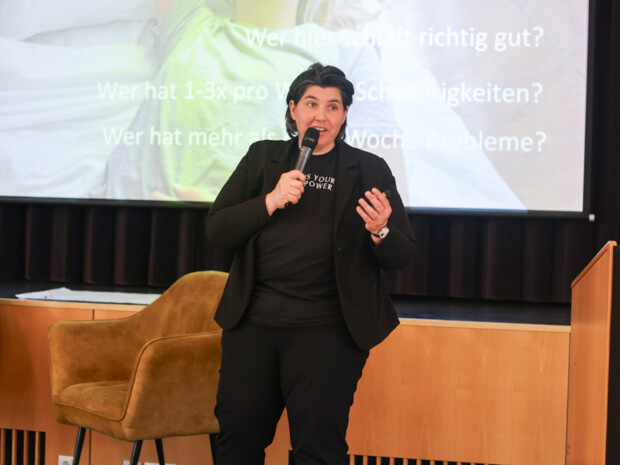 Eine Referentin steht auf der Bühne mit Mikrofon vor einer Präsentationsfolie zum Thema Schlaf und spricht zum Publikum.