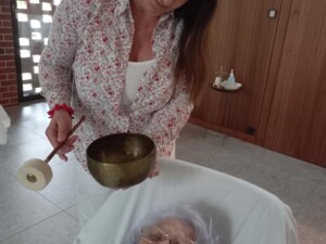 Eine Frau liegt zugedeckt in einem Pflegesessel mit geschlossenen Augen. Eine Betreuerin steht daneben und hält eine Klangschale über ihr - ein Moment der tiefen Ruhe und Entspannung.