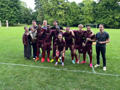 Gruppenfoto eines fröhlichen Fußballteams mit Unterstützer:innen. Die Personen stehen eng beisammen auf einer Wiese, im Hintergrund ist ein Tor und dichter Baumbestand zu sehen.